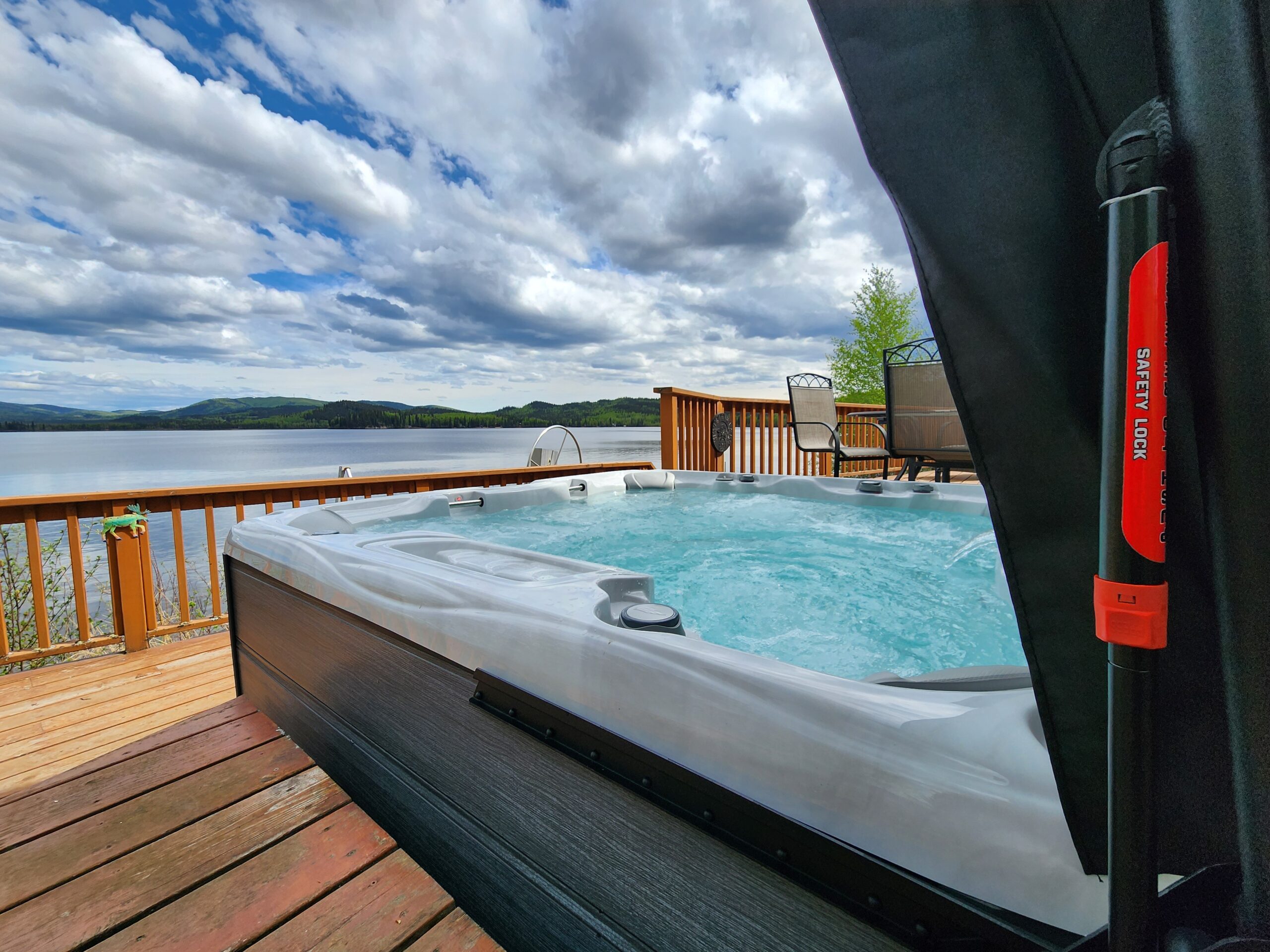 Outdoor hot tub on wooden deck, cover open, close up over covermate hydraulic spring, tub facing lake on a semi cloudy and sunny summer day
